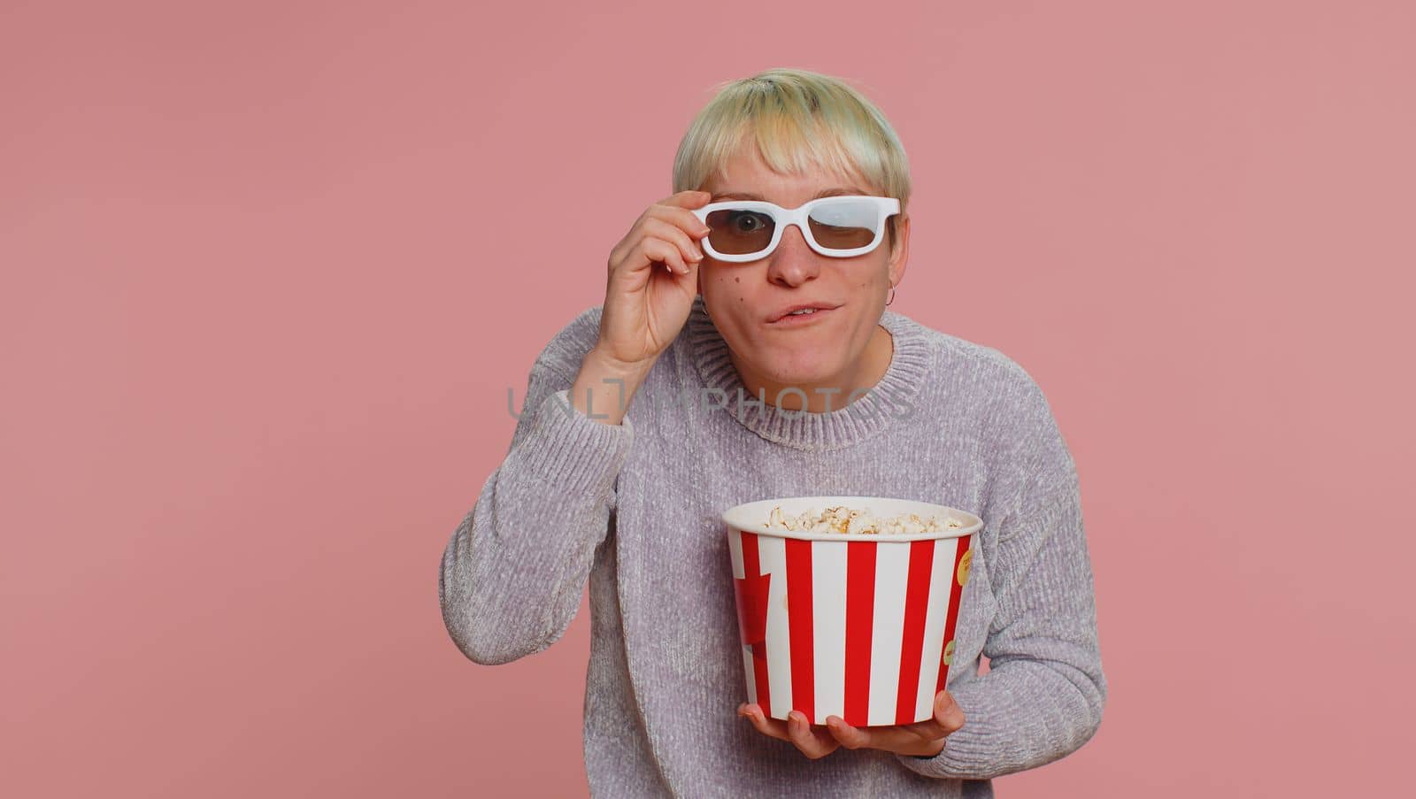 Excited woman with green short hair eating popcorn, watching tv serial, sport game, film, online social media movie content. Young girl in 3D glasses enjoying domestic entertainment on pink background