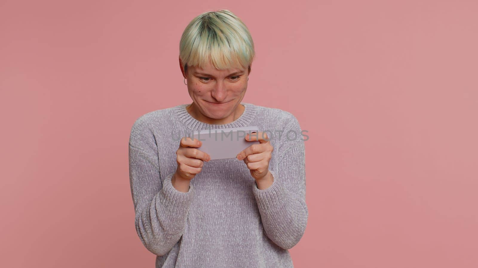 Worried funny addicted woman with short hair enthusiastically playing racing video games on mobile phone. Young girl using smartphone gadget app with drive simulator isolated on pink studio background