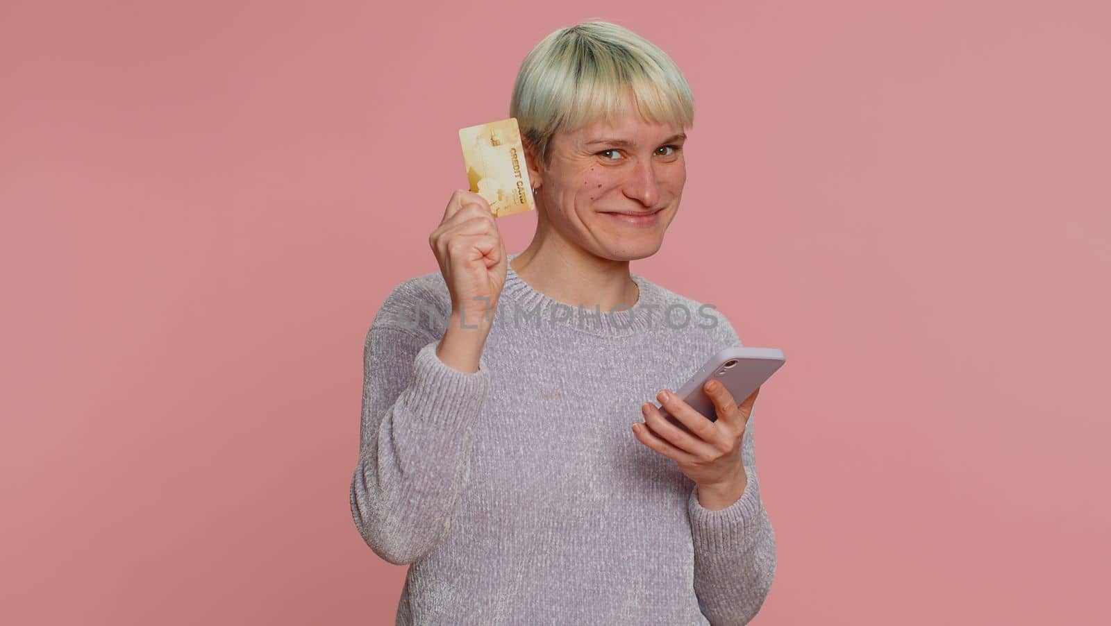 Sincere woman customer using credit bank card and smartphone while transferring money, purchases online shopping, payment. Finance and internet. Young girl on pink background. Lgbt gay lesbian people