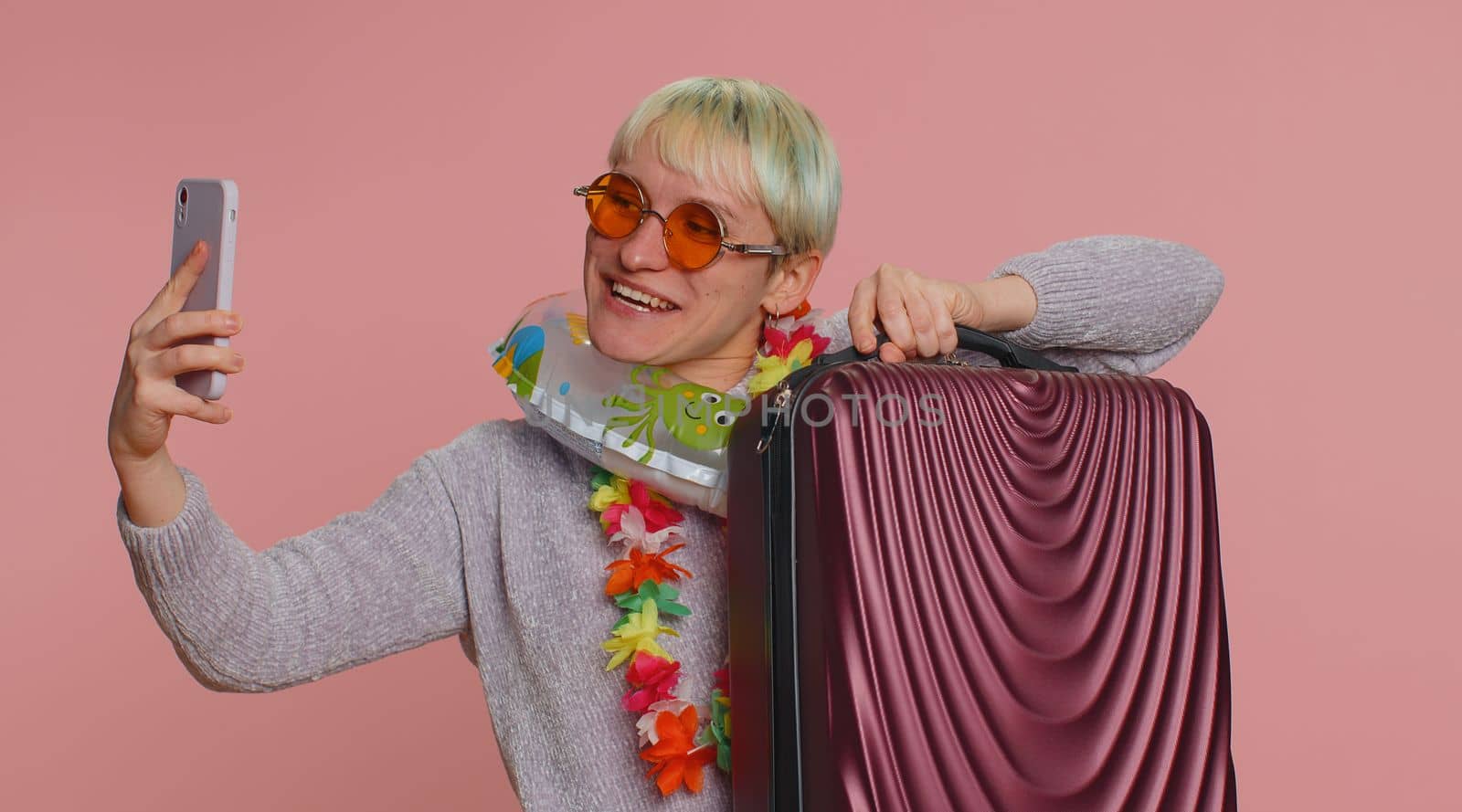 Traveler blogger woman with short hair with swimming ring and luggage, taking selfie on smartphone, make video call online. Young girl on summer holiday vacation, trip. Tourism. Pink studio background