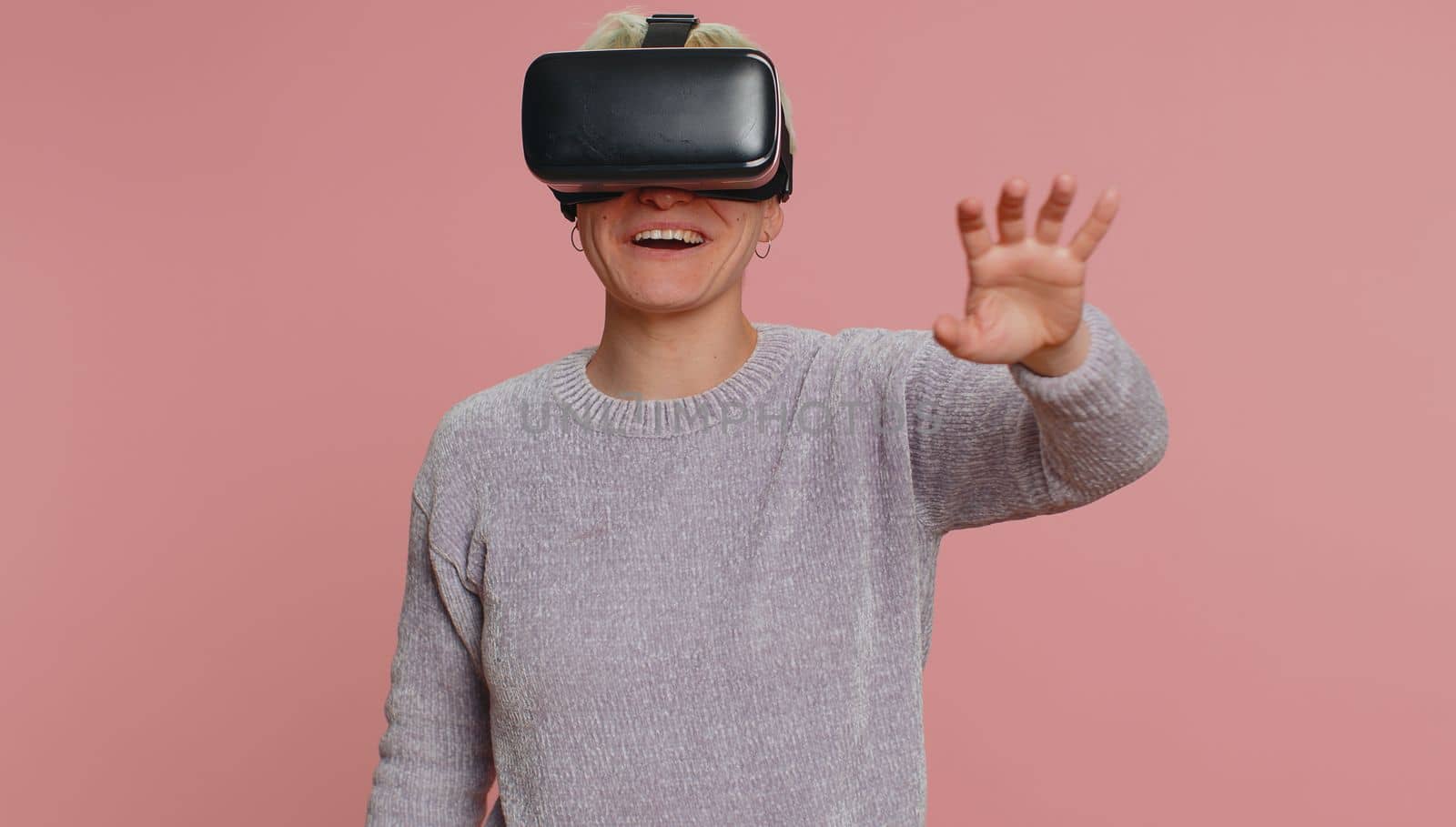 Young woman using headset helmet to play simulation game app online. Watching virtual reality 3D 360 video content. Caucasian one girl in VR goggles on studio pink background. Futuristic technology