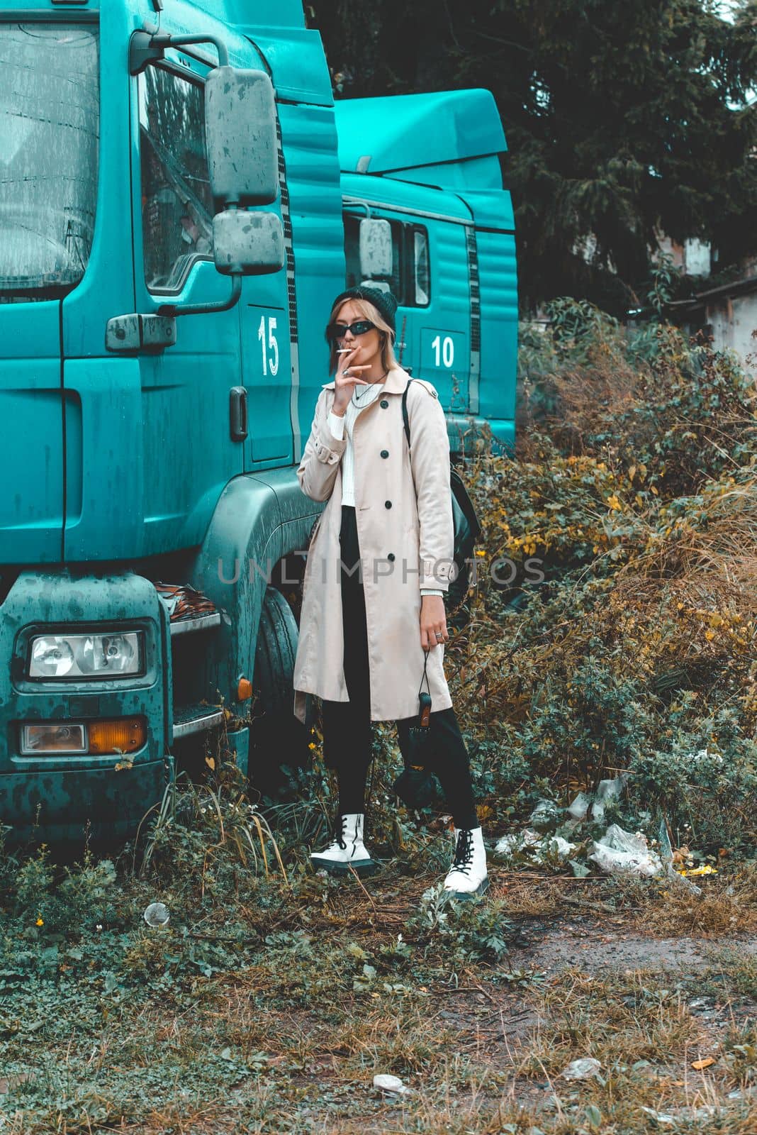 stylish blonde woman wearing long beige coat, white boots and black hat posing near the blue truck. Trendy casual outfit. Selective focus, grain by Ashtray25