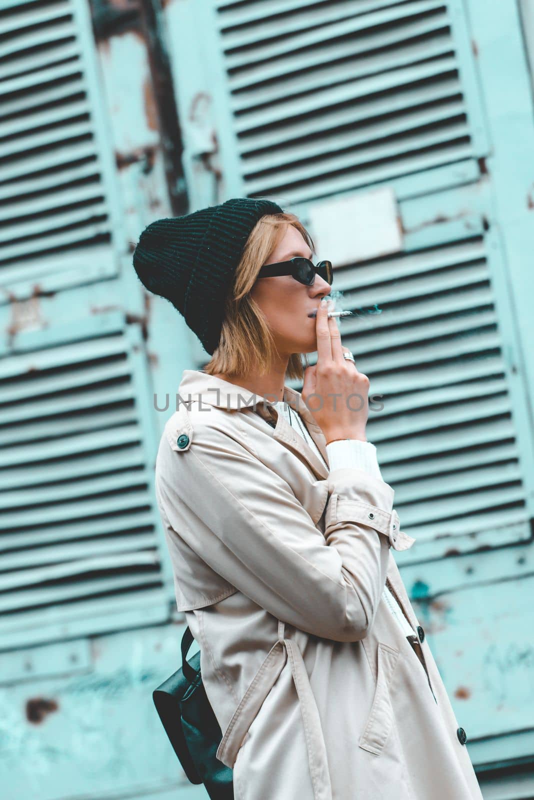 stylish blonde woman wearing long beige coat, white boots, black hat and backpack posing near blue jalousie door. Selective focus, grain by Ashtray25