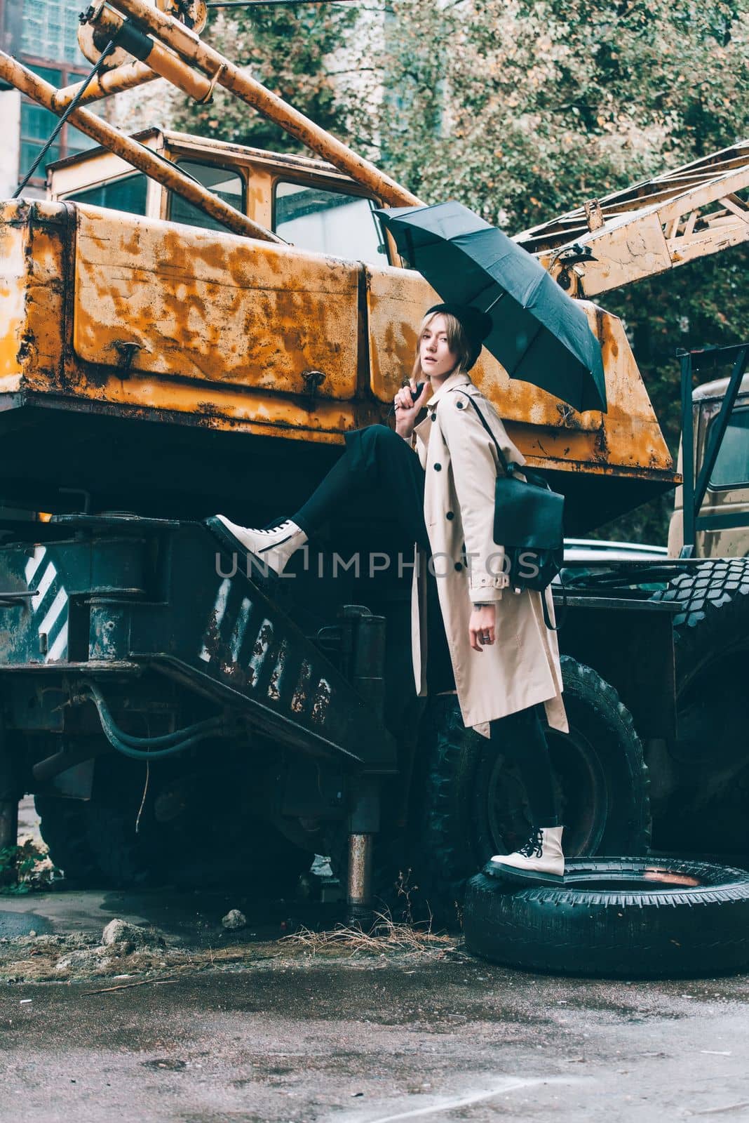 young stylish woman wearing long beige coat, white boots, black hat and umbrella posing near old truck crane. Trendy casual outfit. Selective focus, grain Street fashion. author's toning