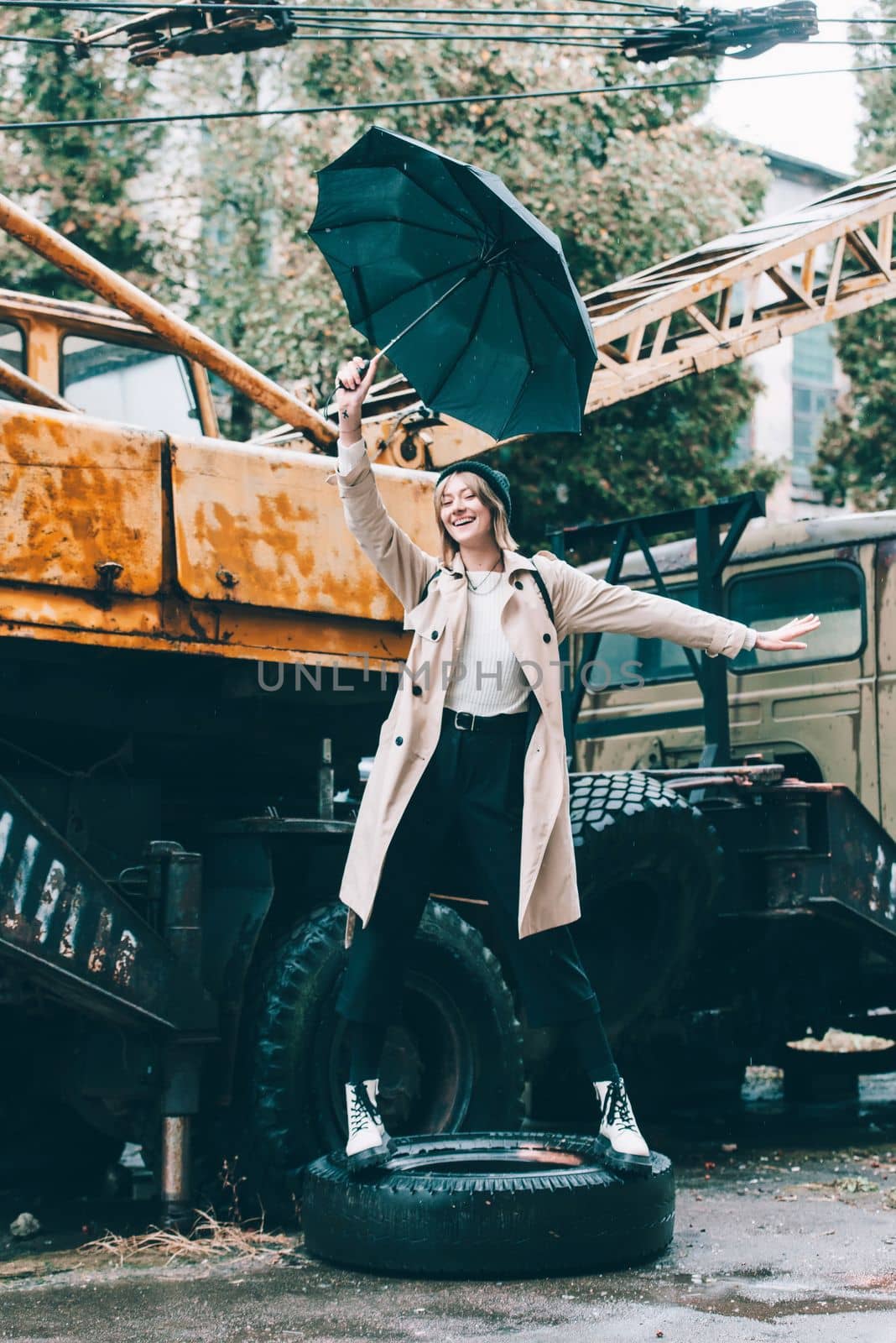 young stylish woman wearing long beige coat, white boots, black hat and backpack posing near truck crane. Trendy casual outfit. Selective focus, grain by Ashtray25