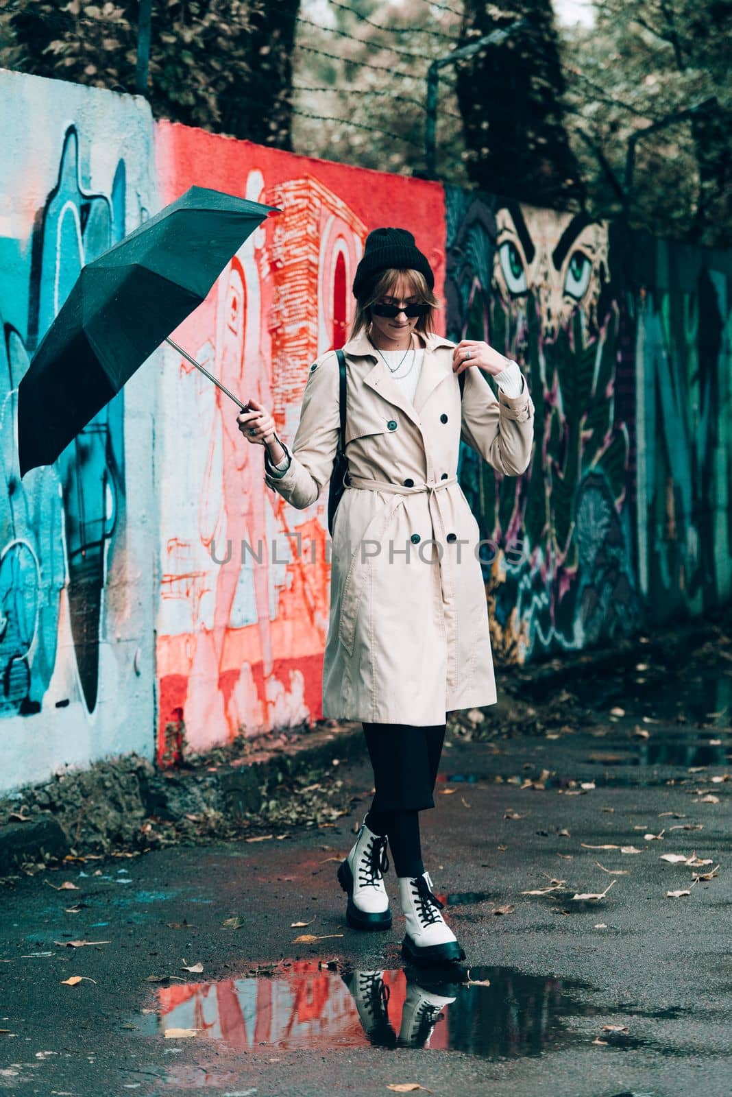Beautiful young stylish blonde woman wearing long beige coat, white boots, black hat, umbrella and backpack posing through the city streets. Trendy casual outfit. Selective focus, grain