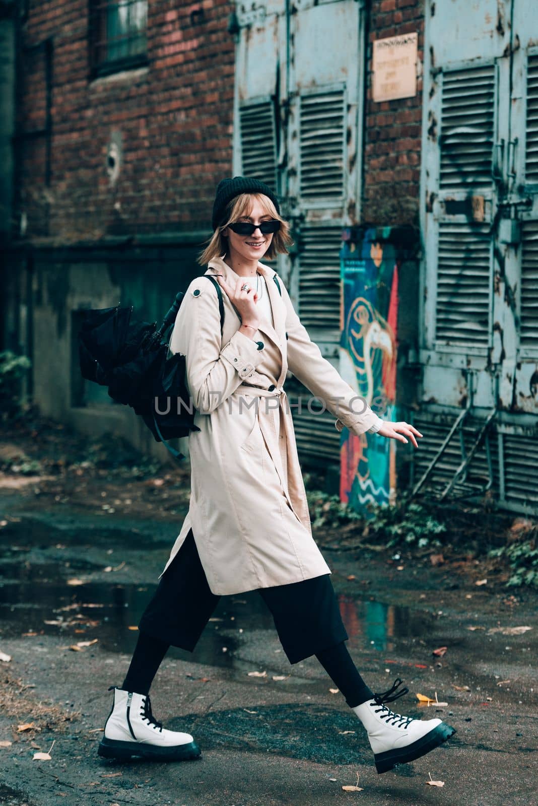 young stylish woman wearing long beige coat, white boots, black hat, umbrella and backpack posing through the city streets. Selective focus, grain by Ashtray25