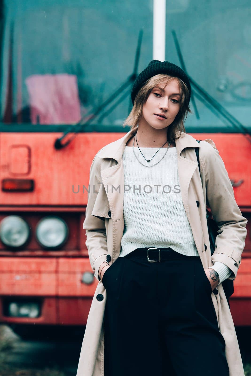 Beautiful young stylish blonde woman wearing long beige coat, white boots and black hat posing on the old red bus. Trendy casual outfit. Selective focus, grain. Street fashion. author's toning