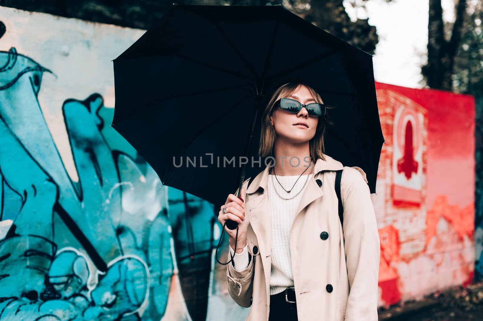 Beautiful young stylish blonde woman wearing long beige coat, white boots, black hat, umbrella and backpack posing through the city streets. Trendy casual outfit. Selective focus, grain