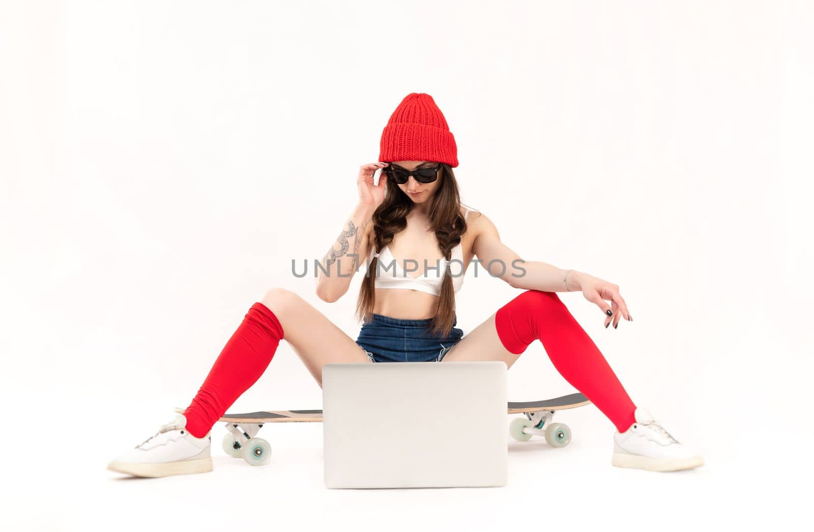 sexy girl in a fashionable red hat is sitting on a longboard on a white background with a laptop