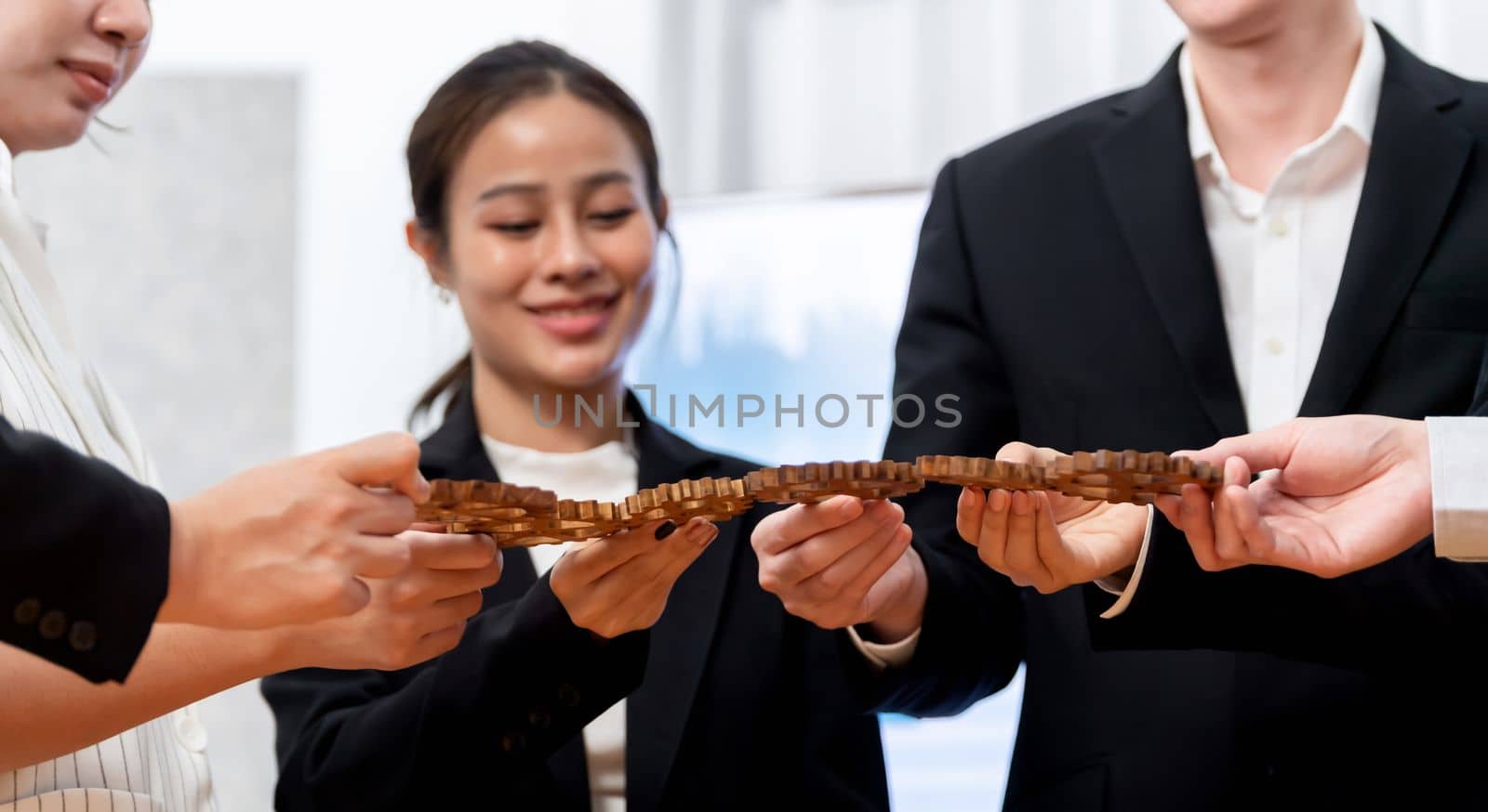 Focus closeup hands holding gears join together in workplace by blur businesspeople in background as concept of harmony in office. Synergy by office workers making chain cogwheel into collective form.