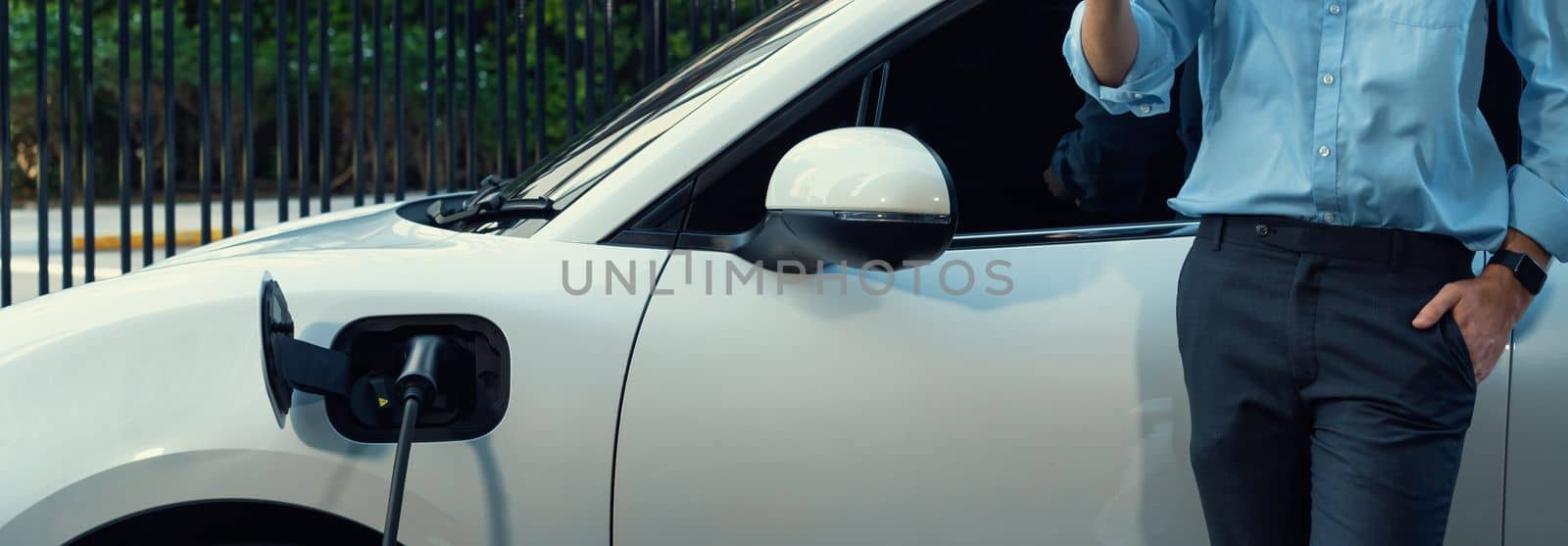 Closeup progressive suit-clad businessman with his electric vehicle recharge his car on public charging station in modern city with power cable plug and renewable energy-powered electric vehicle.