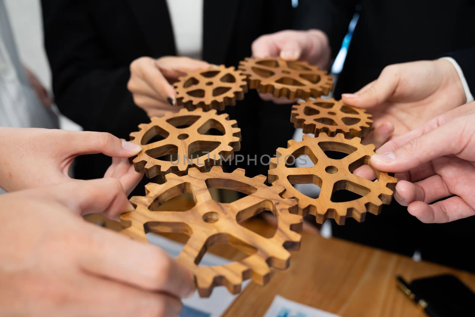 Closeup top view businesspeople hand holding gear and join together over meeting table with financial report papers. Cohesive group of office workers holding cog wheel as synergy harmony concept.