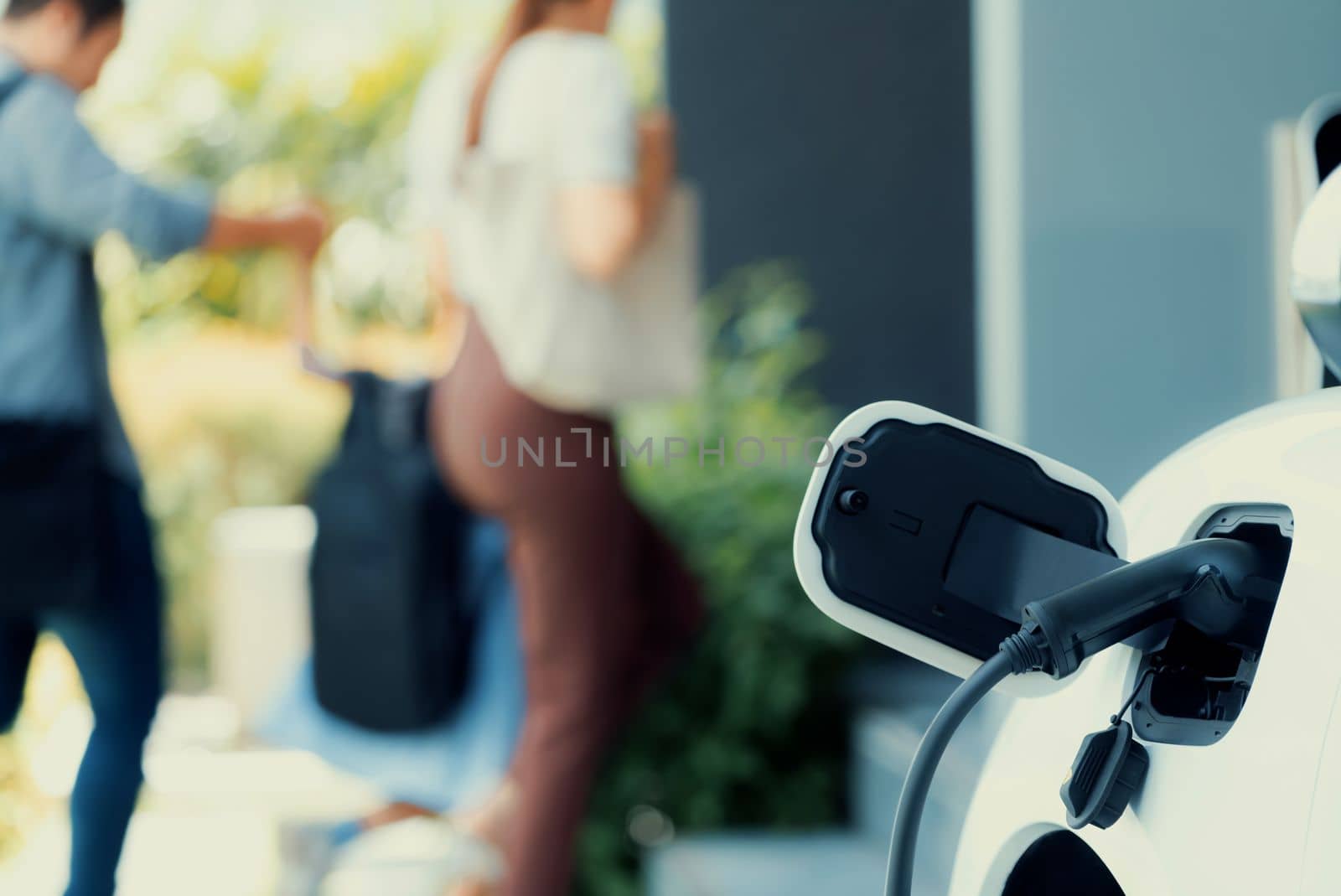 Focus closeup electric vehicle recharging battery from home electric charging station with blurred family in background. Renewable clean energy car for progressive eco awareness lifestyle concept.