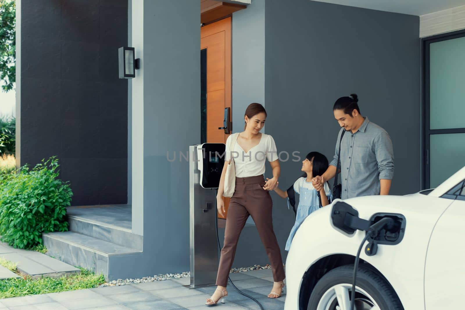 Progressive young parents and daughter with electric vehicle and home charging station. Green and clean energy from electric vehicles for healthy environment. Eco power from renewable source at home.