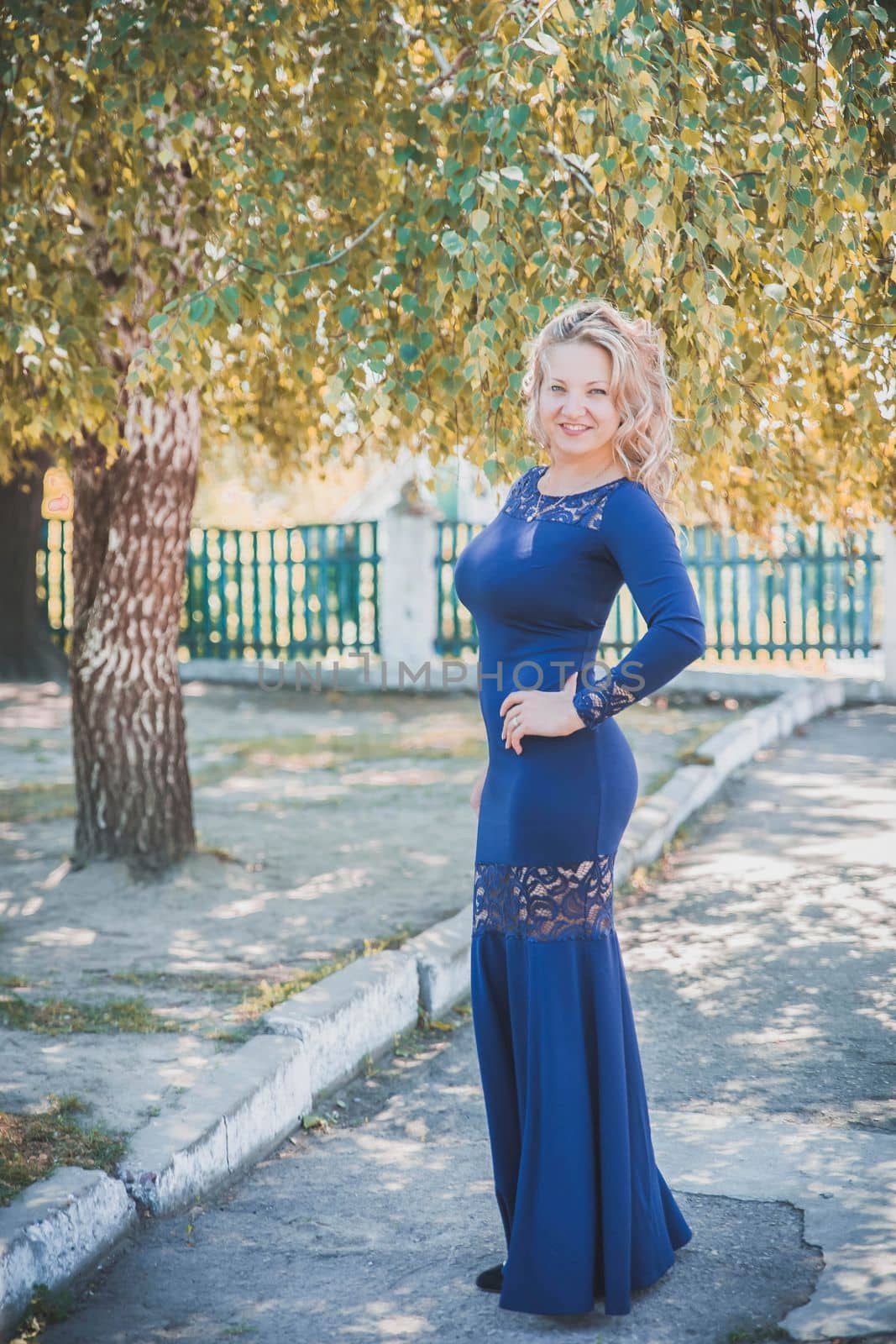 beautiful slim blonde with a long blue dress smiles under autumn tree.