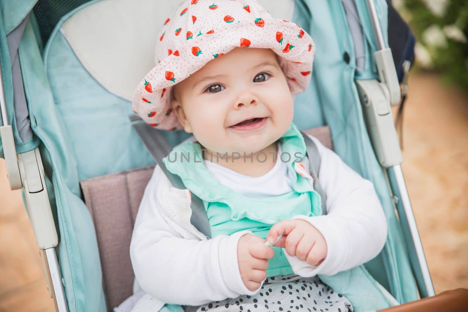 baby in hat smiles toothlessly sitting in a blue stroller.