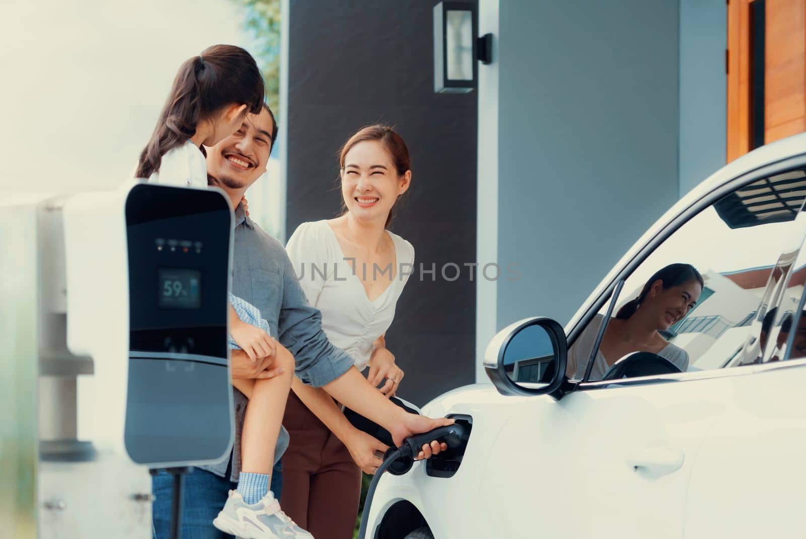 Progressive young parents and daughter with electric vehicle and home charging station. Green and clean energy from electric vehicles for healthy environment. Eco power from renewable source at home.