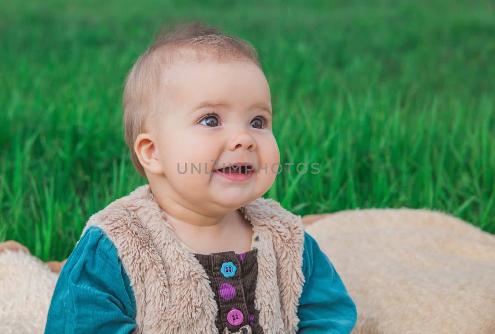 toothless baby in a multi-colored dress sitting on a bedspread on lawn.