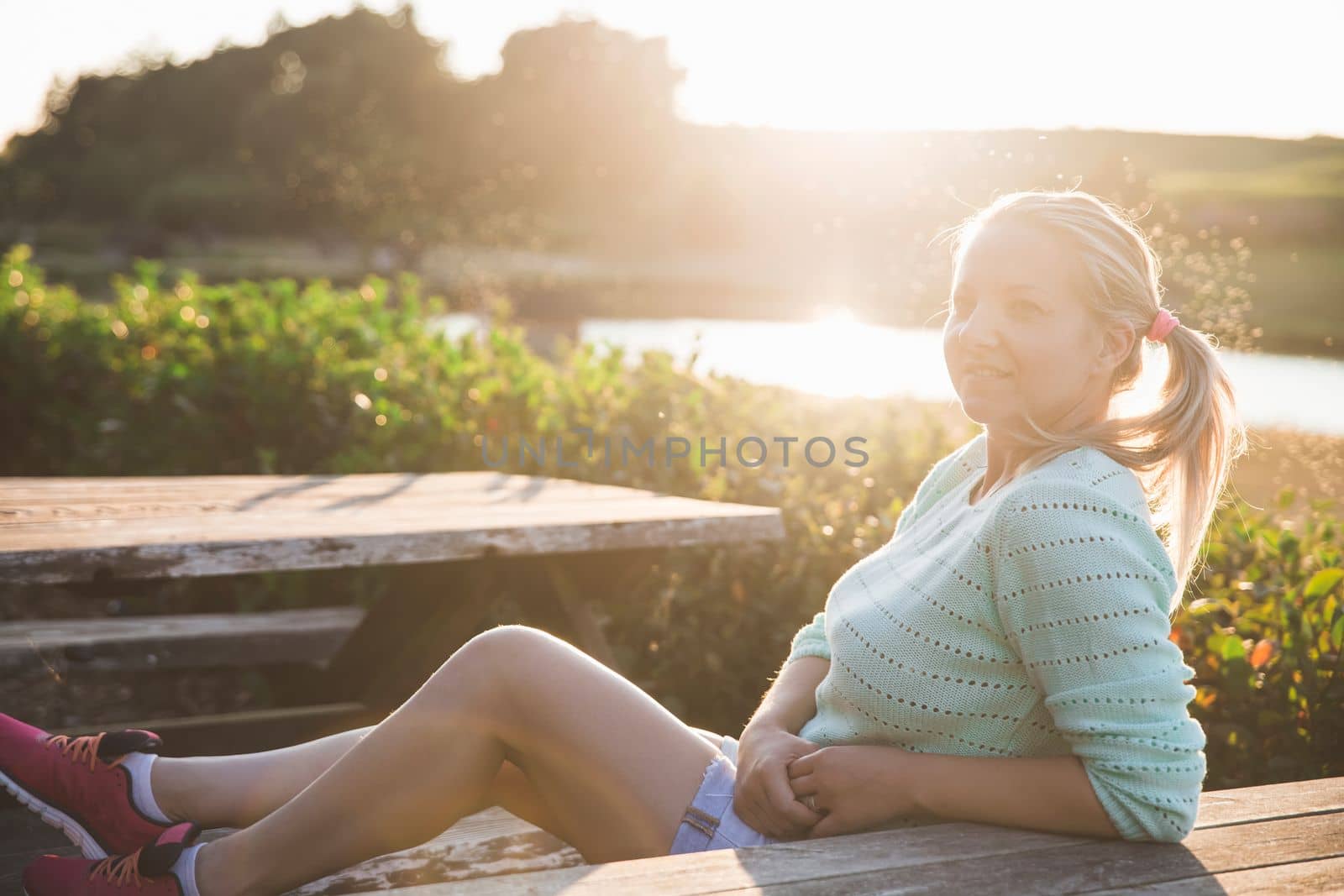 blonde girl in shorts sitting at sunset at the wooden table near the lake