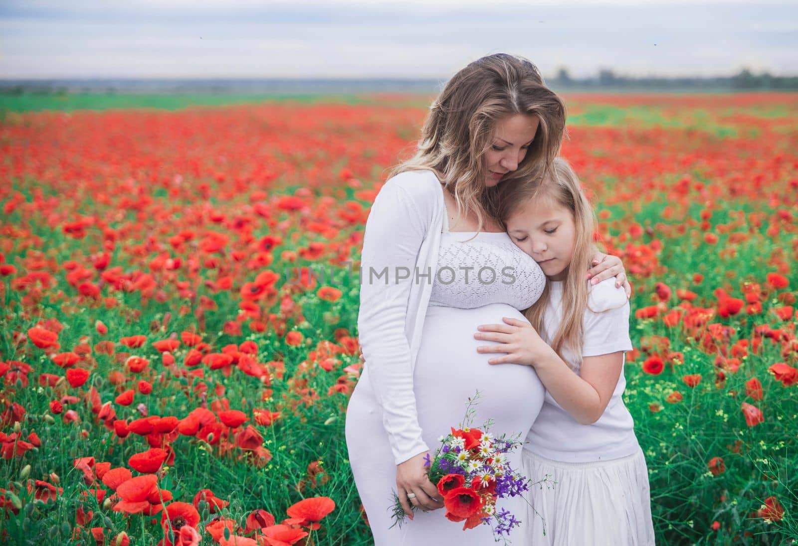 pregnant mother with her eldest daughter dressed in white dresses is cuddle in the poppy field