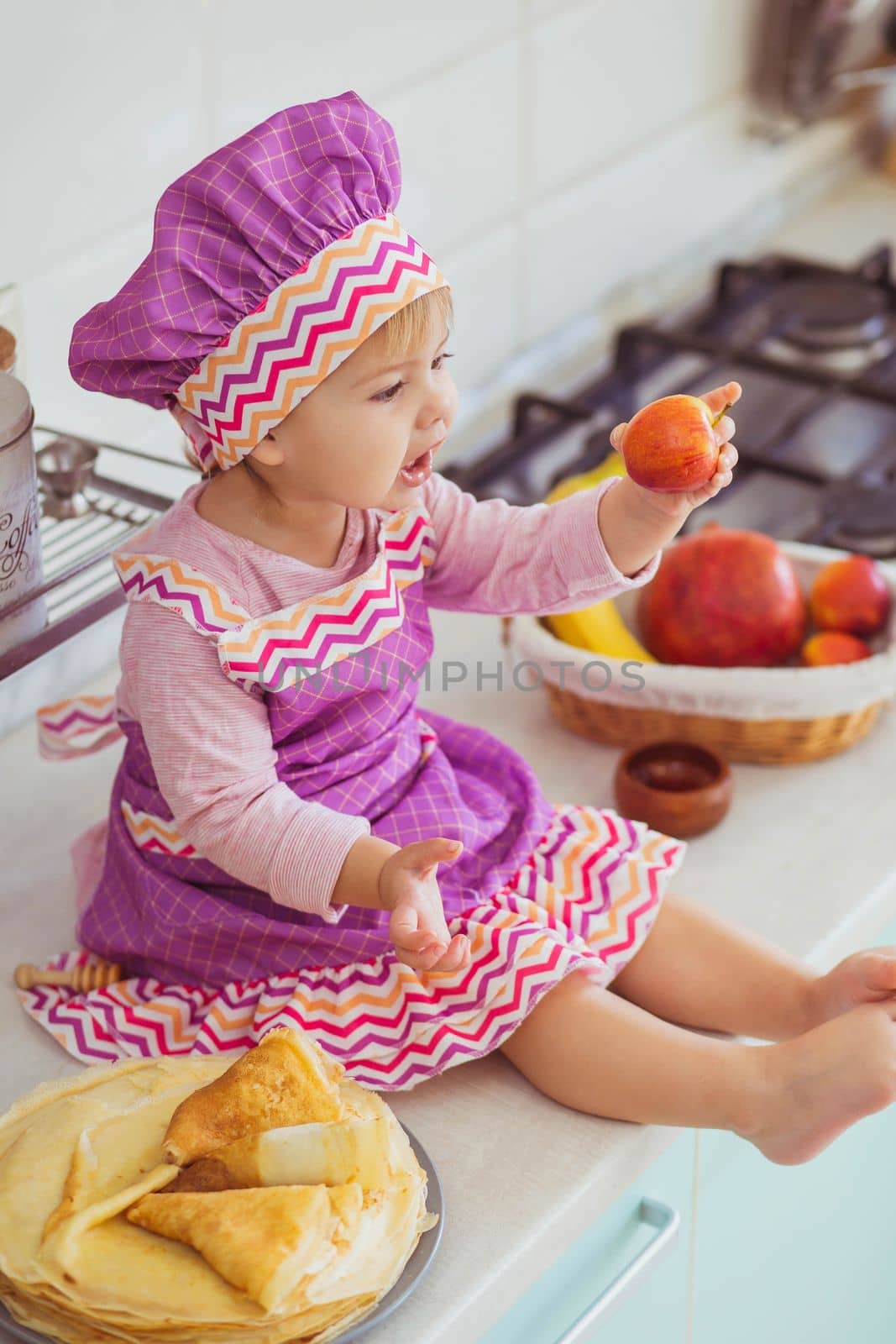 Adorable baby in an apron and chef's cap sitting with an apple on the table.