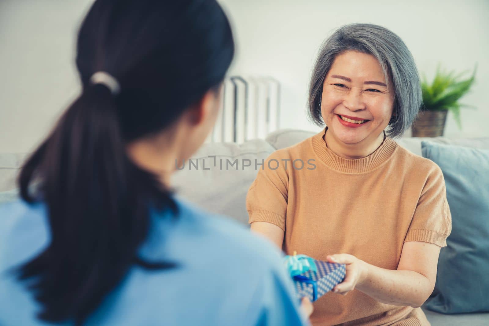 A young caregiver hand over to her senior patient a blue gift box with blue ribbons at a contented living room.