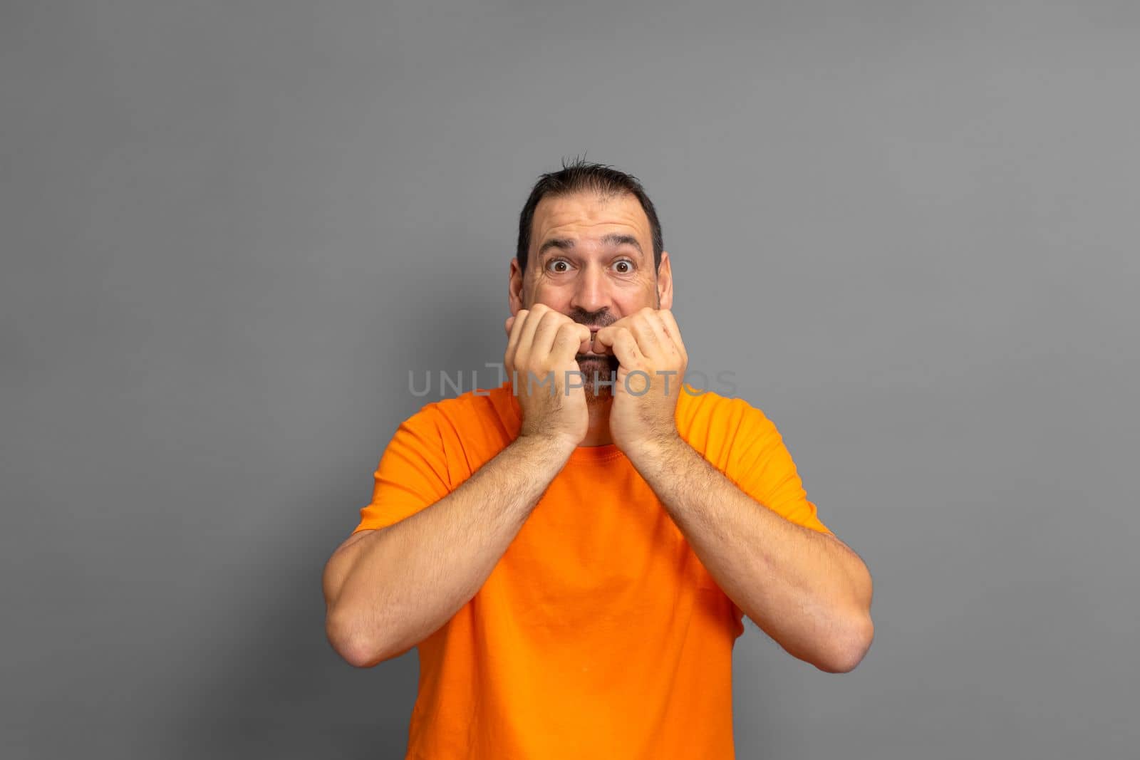 Latino man with a beard in an orange t-shirt biting his nails out of fear, he is experiencing a distressing situation. Isolated on gray background