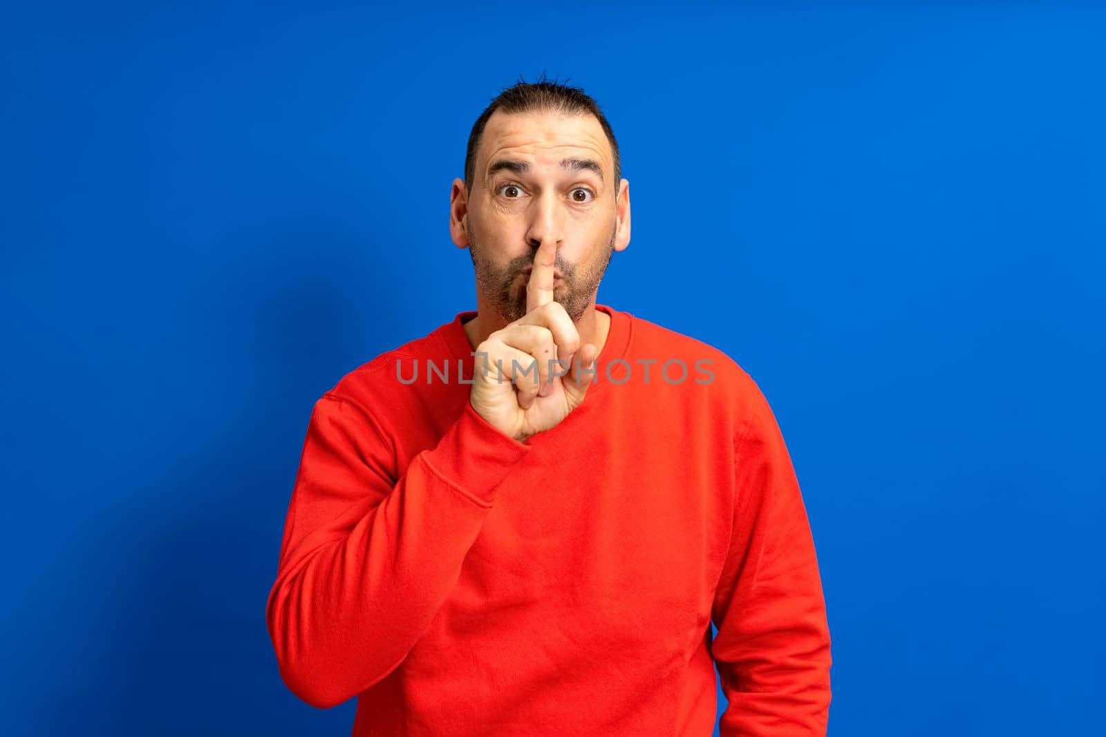 Serious handsome bearded hispanic man puts his index finger to his lips, tries to keep conspiracy, looks aside and says: Shh, be quiet please. Isolated shot of attractive man shows silence sign