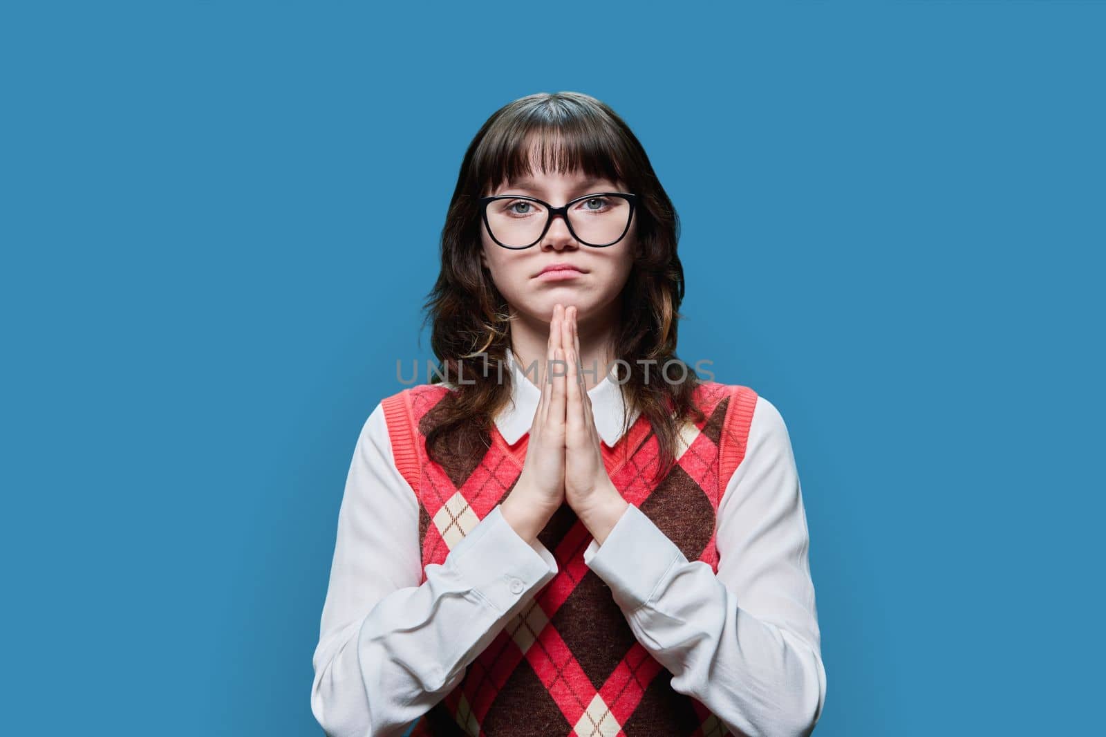 Young sad female in glasses pleading holding her palms together, on blue studio background. Girl student looking at camera, in request, in plea, asking for forgiveness for help sorry pray please