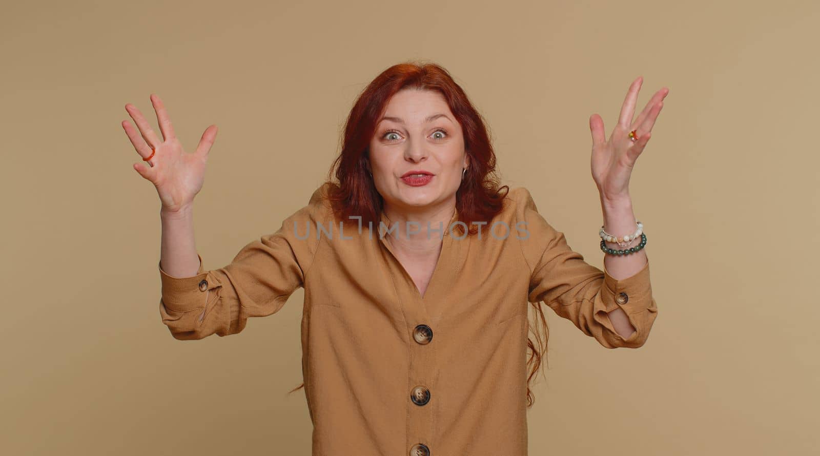 Excited amazed redhead woman in blouse touching head and showing explosion, looking worried and shocked, professional burnout. Looking surprised wow girl isolated alone on beige studio wall background