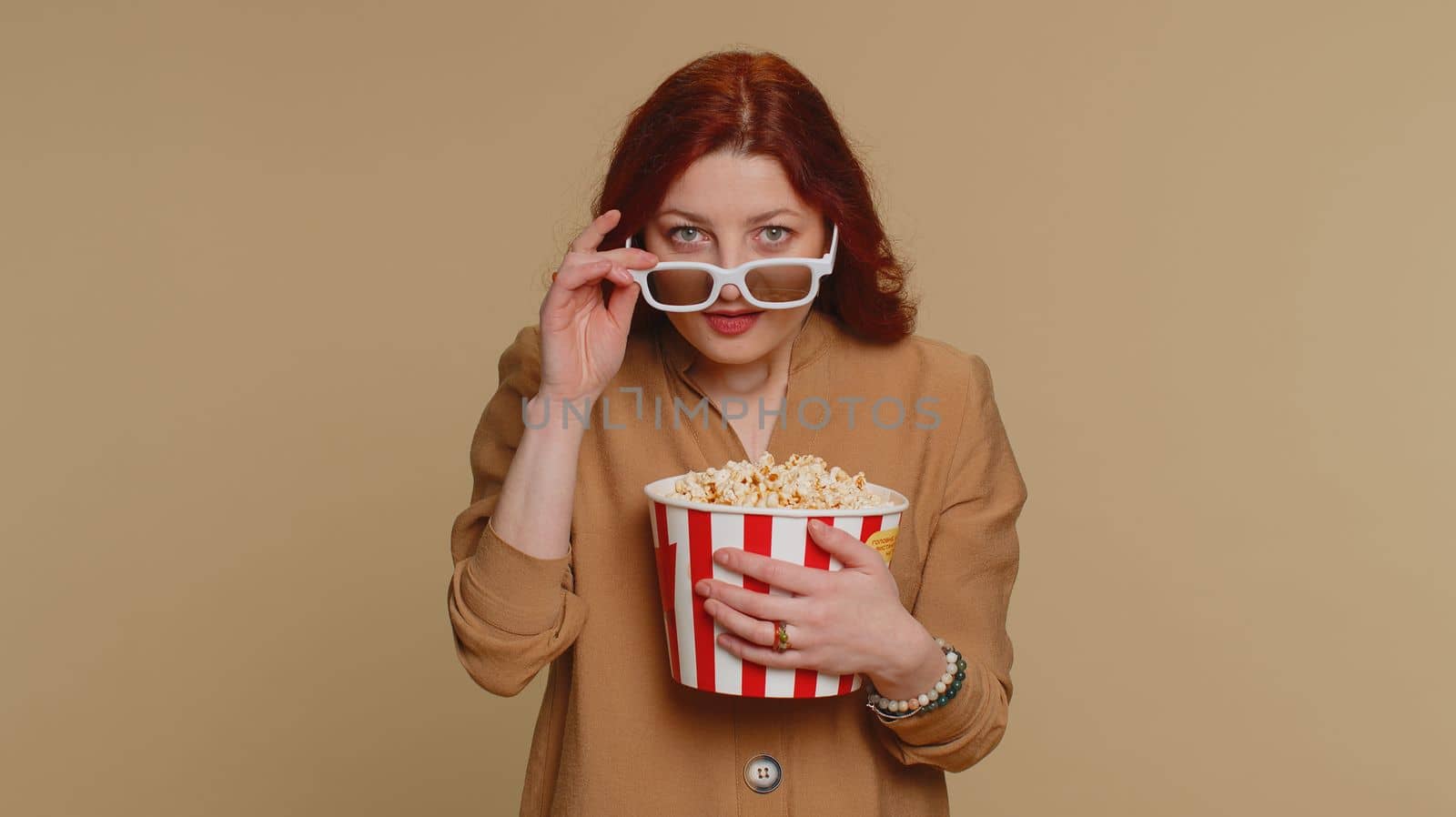 Excited redhead woman in 3D glasses eating popcorn and watching interesting tv serial, sport game, film, online social media movie content. Girl enjoying domestic entertainment on beige background