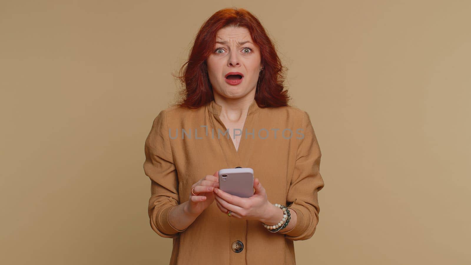 Hipster redhead woman use mobile cell phone typing browsing, loses becoming surprised sudden lottery results, bad fortune, loss, fail. Young girl isolated alone on beige studio wall background