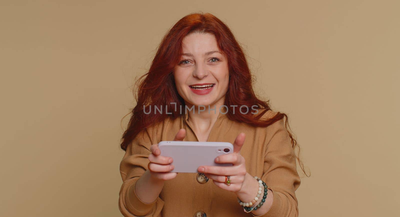 Worried funny addicted woman enthusiastically playing racing video games on mobile phone. Young redhead girl using smartphone gadget app with drive simulator isolated on beige studio background