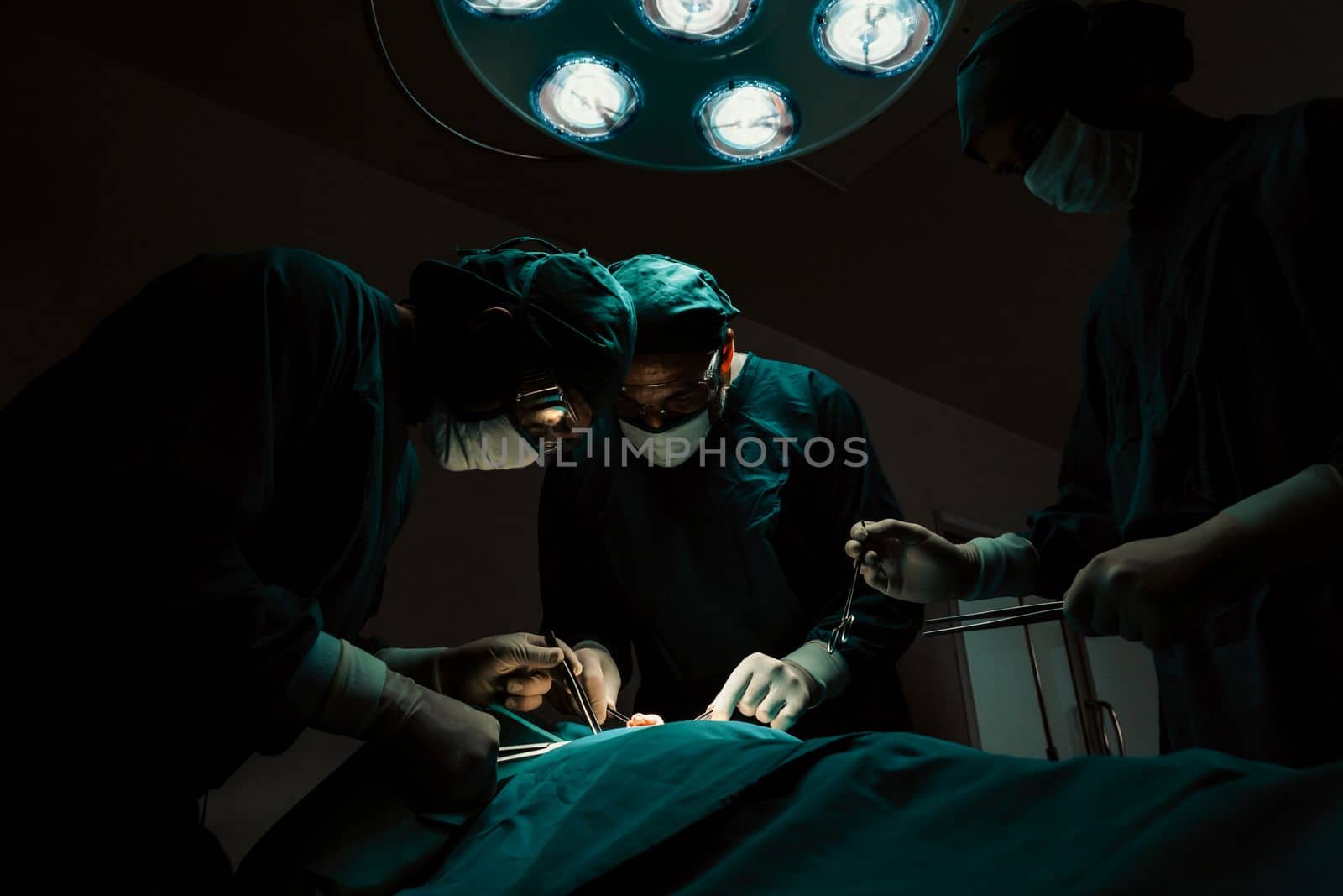 Surgical team performing surgery to patient in sterile operating room. In a surgery room lit by a lamp, a professional and confident surgical team provides medical care to an unconscious patient.