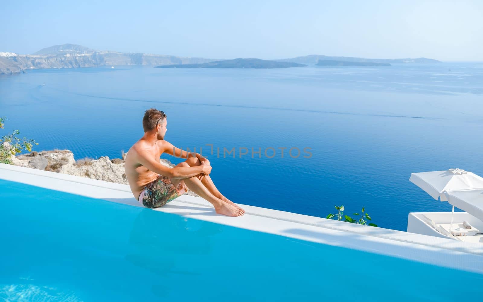 young man relaxing in infinity swimming during vacation at Santorini, swimming pool looking out over the Caldera ocean of Santorini, Oia Greece, Greek Island Aegean Cyclades luxury vacation.