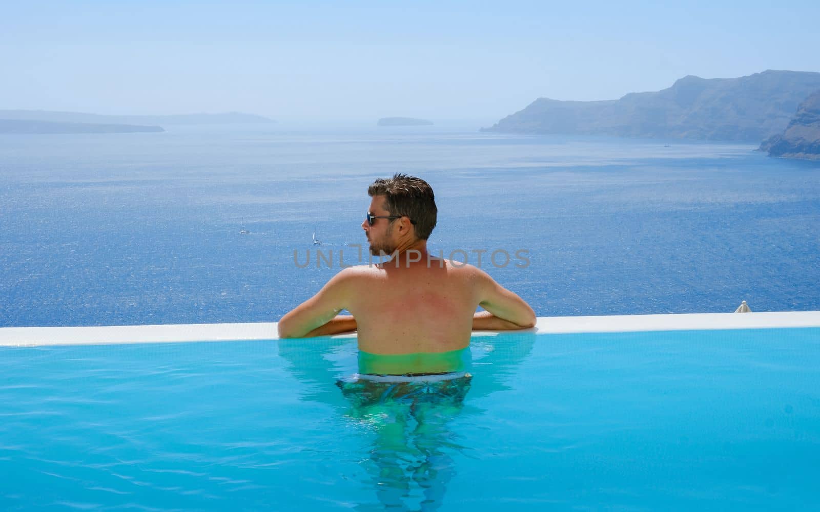 young man relaxing in infinity swimming during vacation at Santorini, swimming pool looking out over the Caldera ocean of Santorini, Oia Greece, Greek Island Aegean Cyclades luxury vacation.