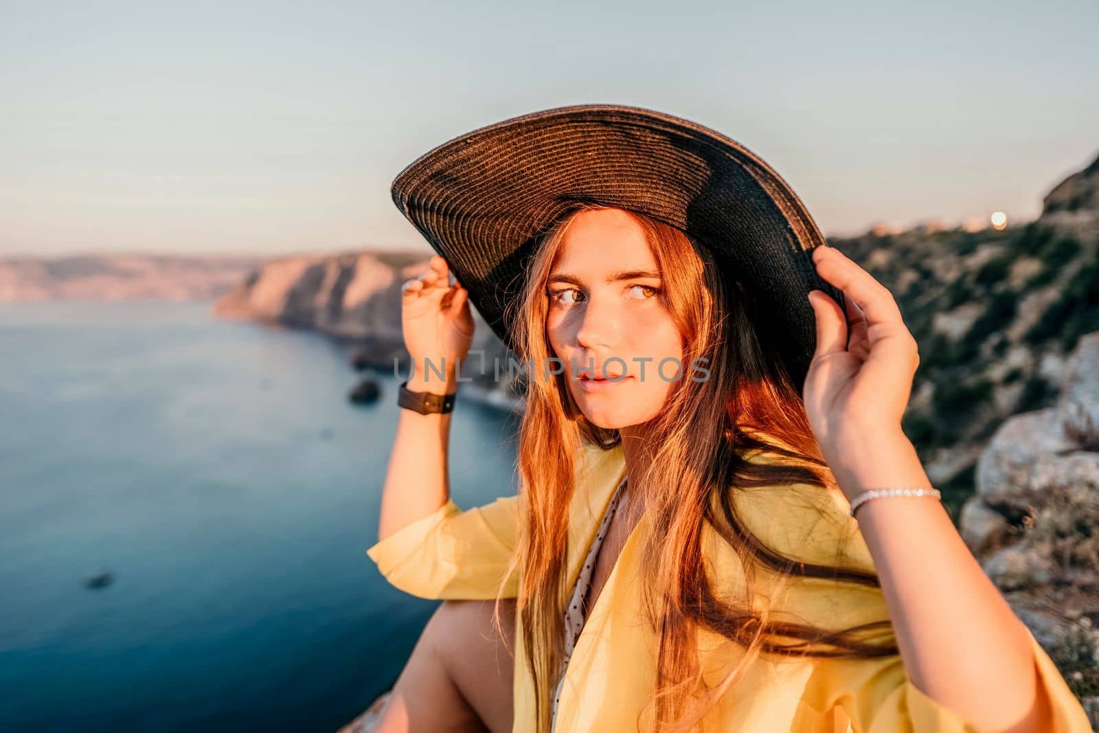 Portrait of happy young woman wearing summer black hat with large brim at beach on sunset. Closeup face of attractive girl with black straw hat. Happy young woman smiling and looking at camera at sea