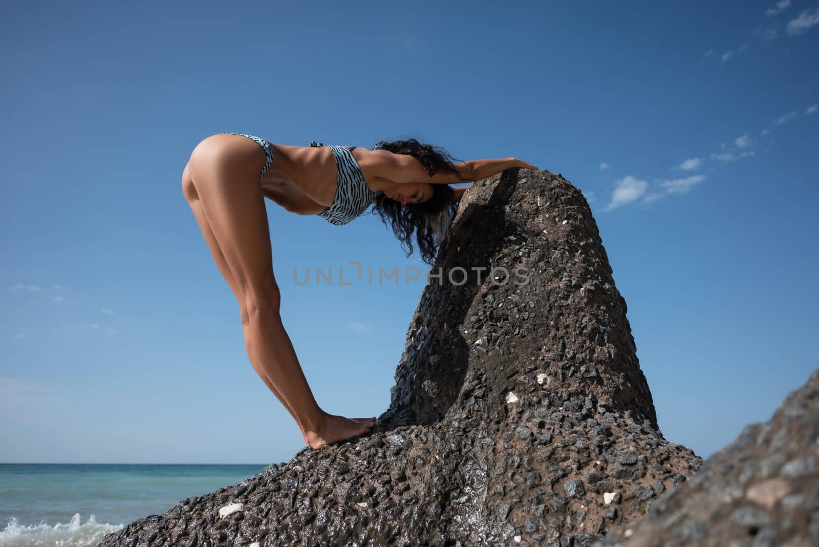 sexy yoga girl in a swimsuit poses slimly on a rock against the background of the sea by Rotozey