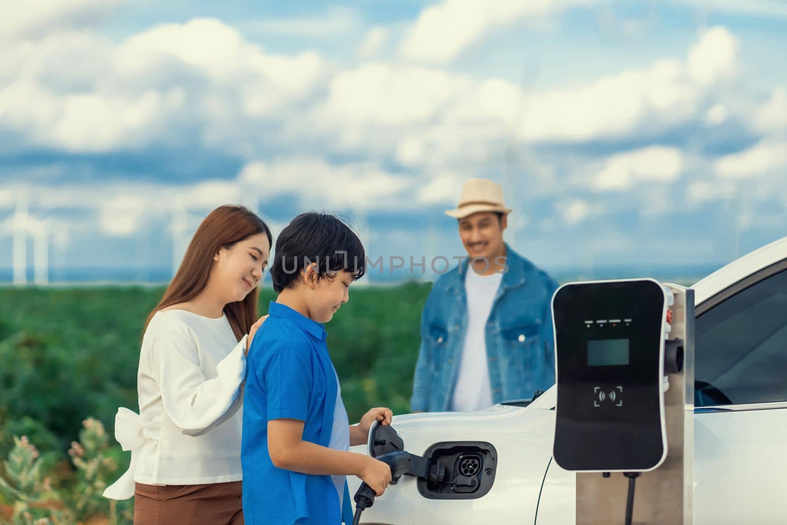 Concept of progressive happy family enjoying their time at wind farm with electric vehicle. Electric vehicle driven by clean renewable energy from wind turbine generator for charging station.