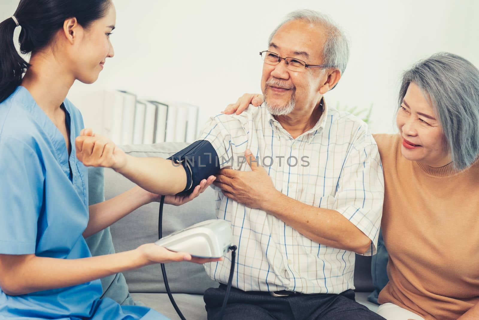 An elderly man having a blood pressure check by his personal caregiver with his wife sitting next to him in their home.
