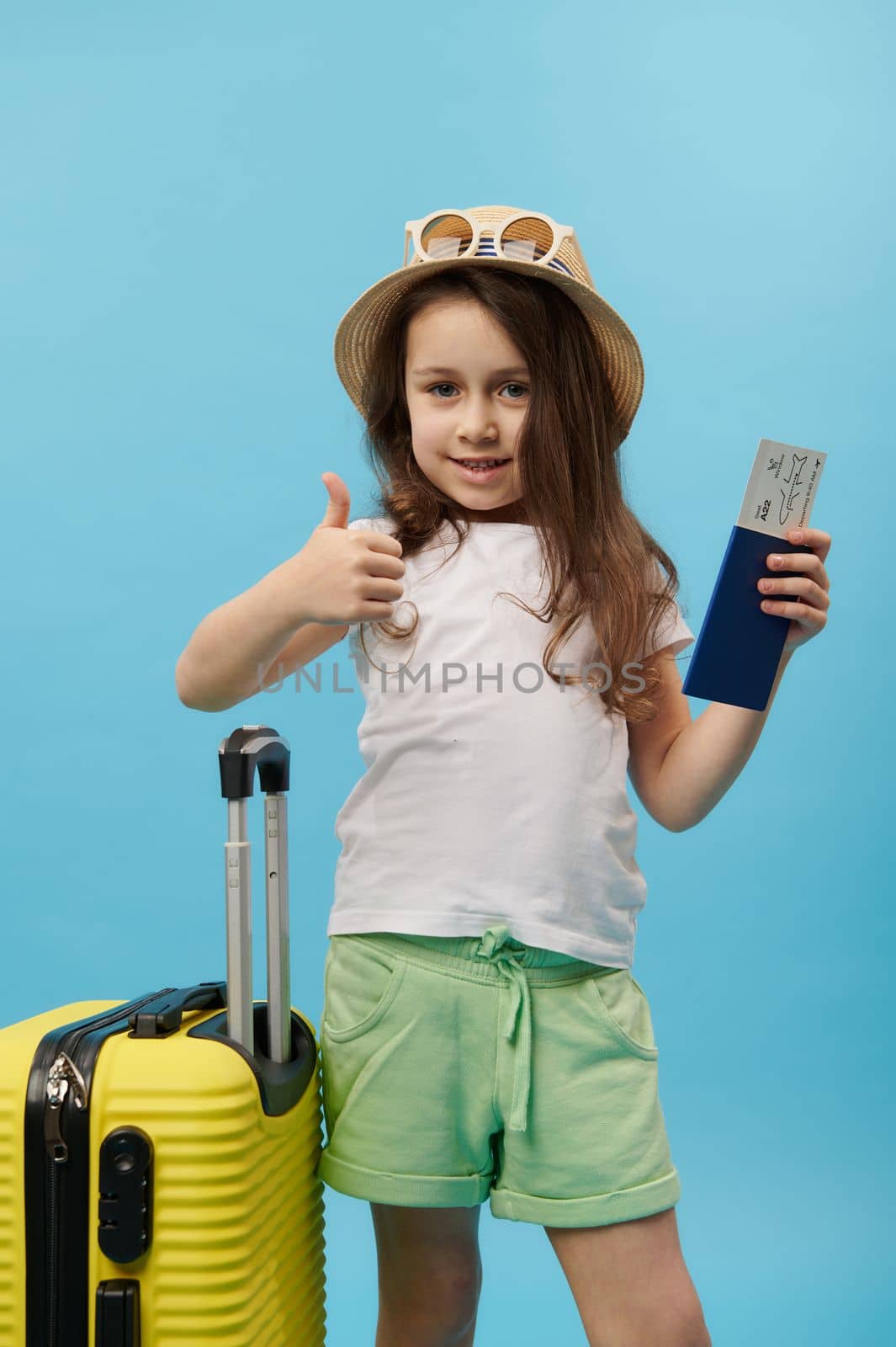 Adorable lovely 5 years little traveler girl in summer wear, showing thumb up looking at camera, posing with a flight ticket , boarding pass and yellow suitcase, blue color background. Trip and travel