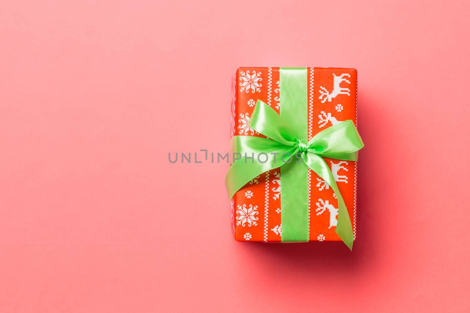wrapped Christmas or other holiday handmade present in paper with green ribbon on living coral background. Present box, decoration of gift on colored table, top view with copy space.