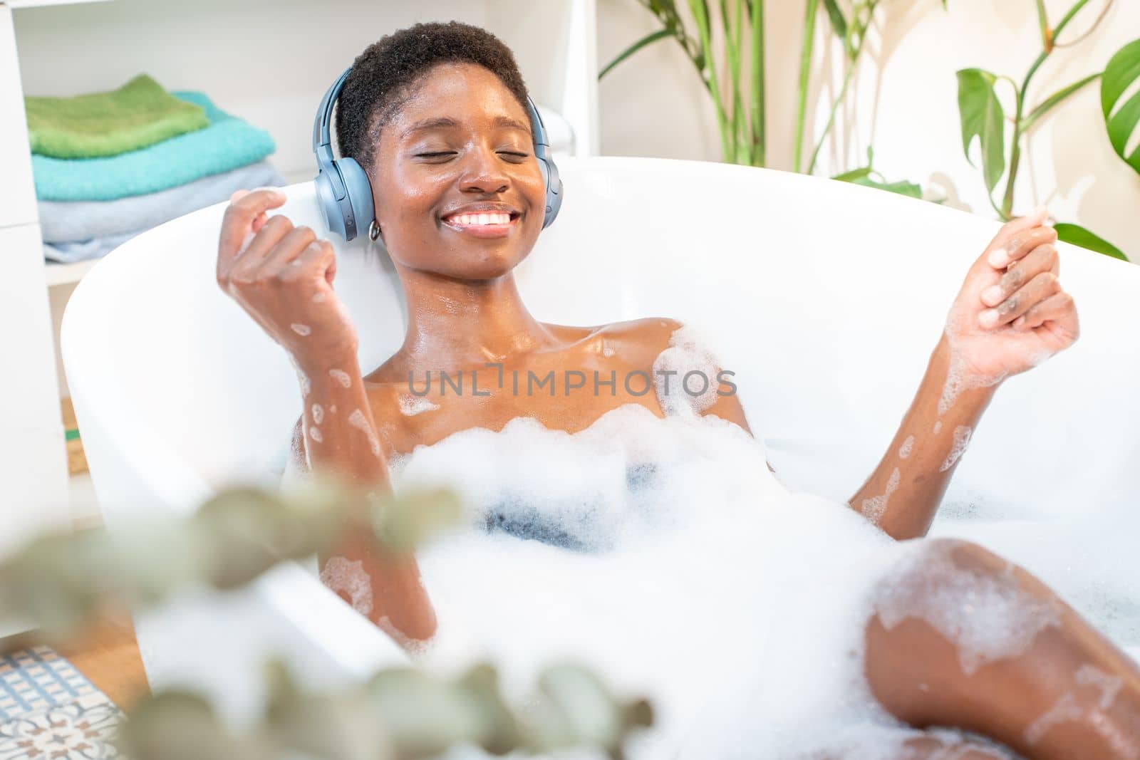 Attractive black woman wearing headphones happy relaxing and dancing in foam bath in beautiful bathroom with plants. Beauty, skin care and wellbeing concept. High quality photo