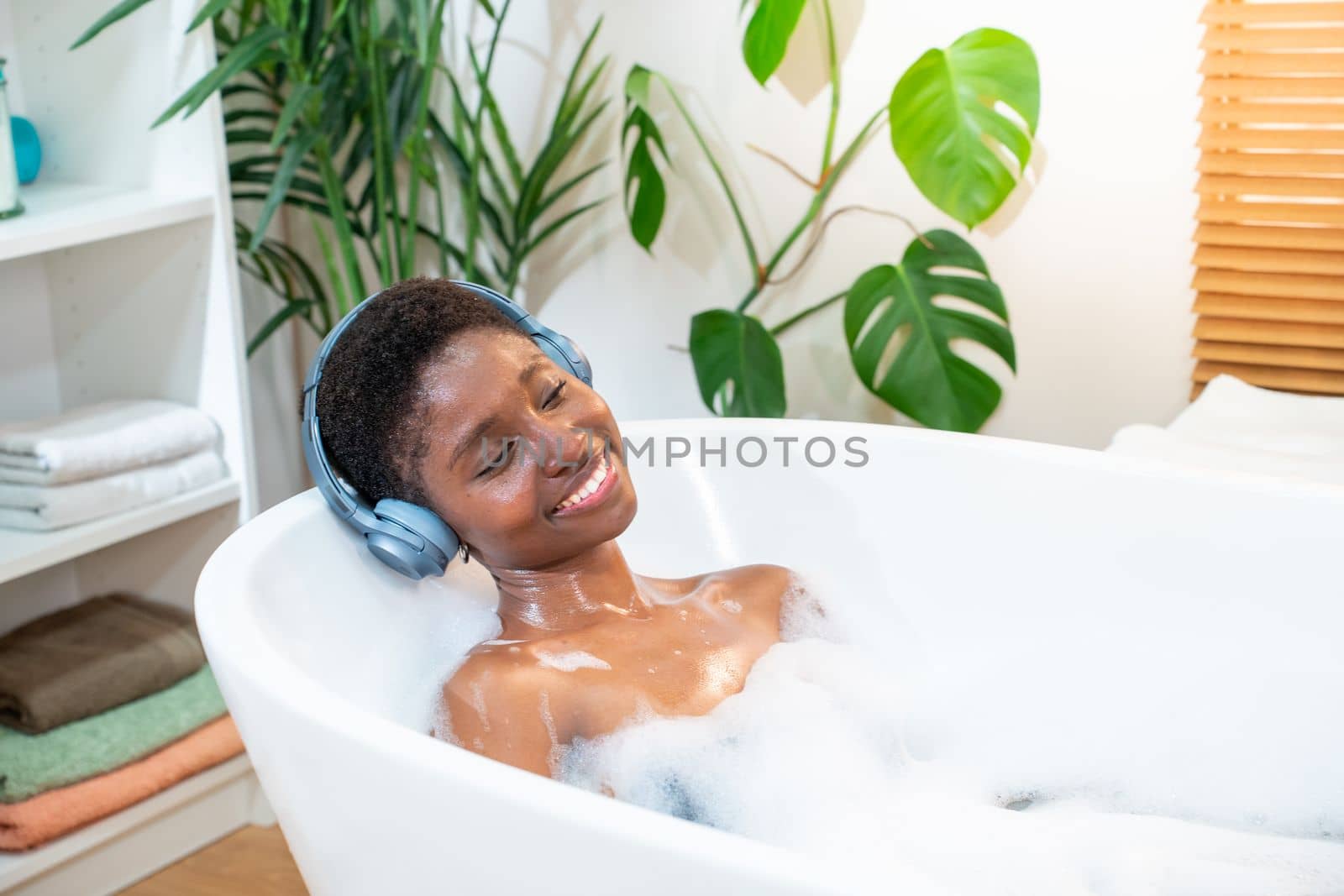 Smiling African attractive woman wearing headphones relaxing and dancing in foam bath in bathroom. by PaulCarr