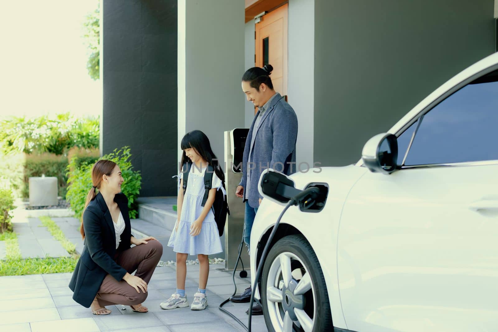 Progressive young parents and daughter with electric vehicle and home charging station. Green and clean energy from electric vehicles for healthy environment. Eco power from renewable source at home.