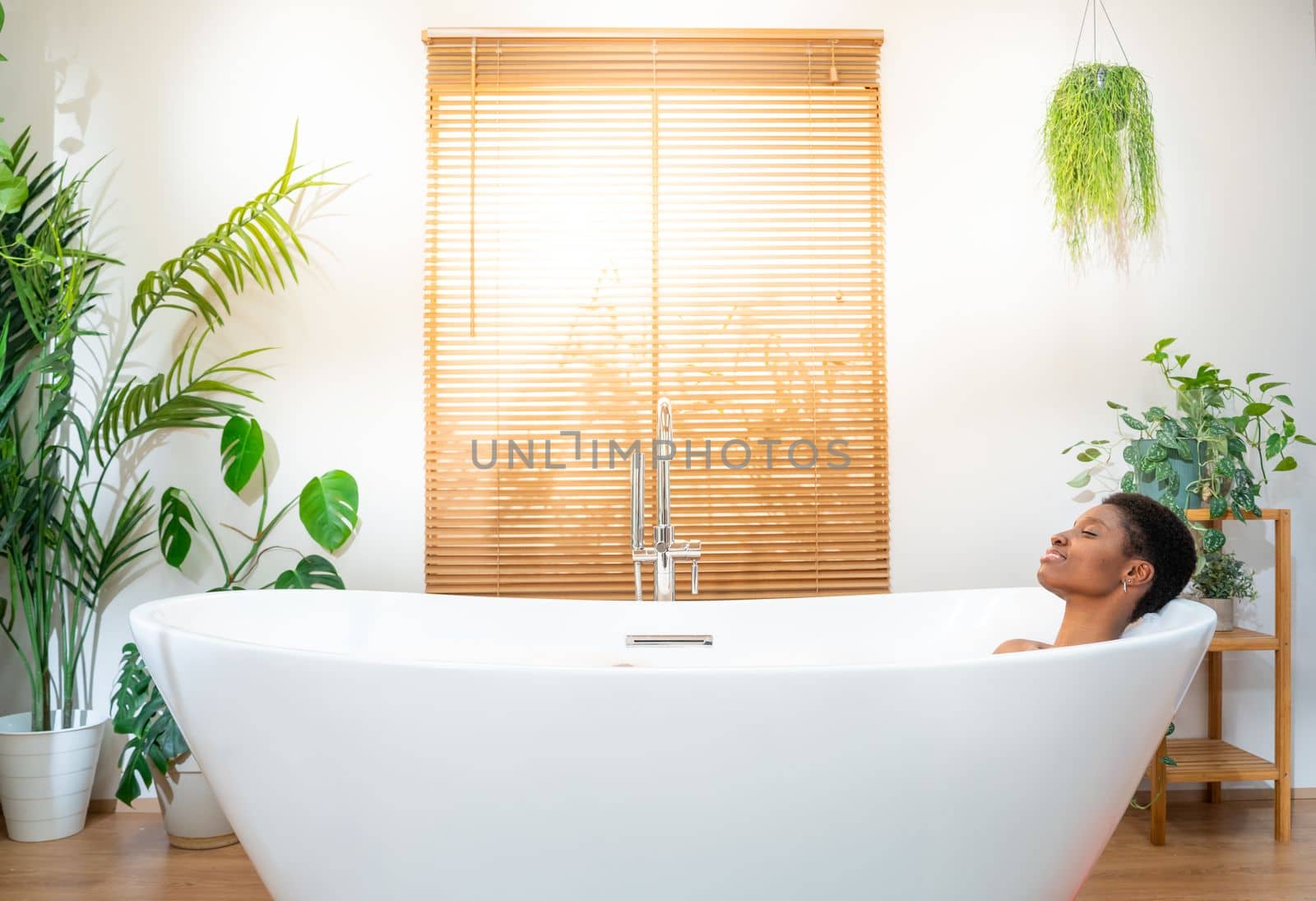 Attractive woman relaxing in foam bath in beautiful bathroom with plants at home during a sunny morning routine. Rest, calm, pleasure, happiness and wellbeing concept. High quality photo