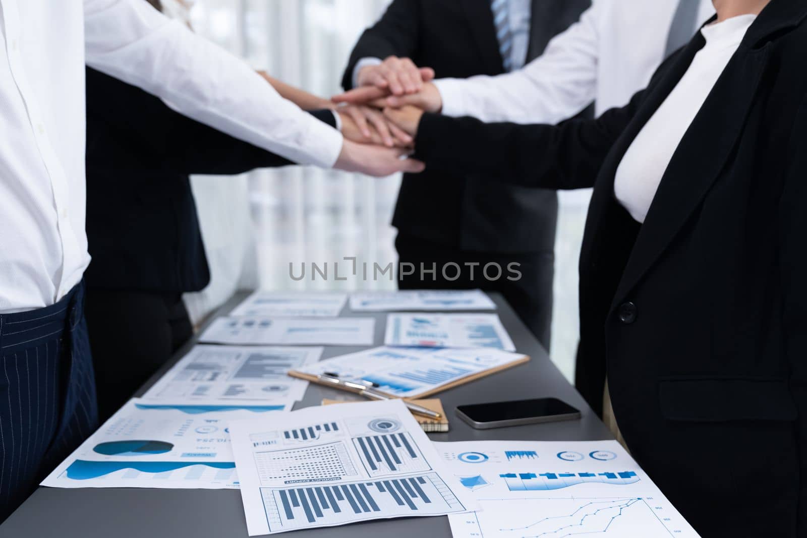 Focus financial report with statistic charts analyzed by BI papers on meeting table at conference room with blur business colleague people put hand stack together in background as harmony concept.