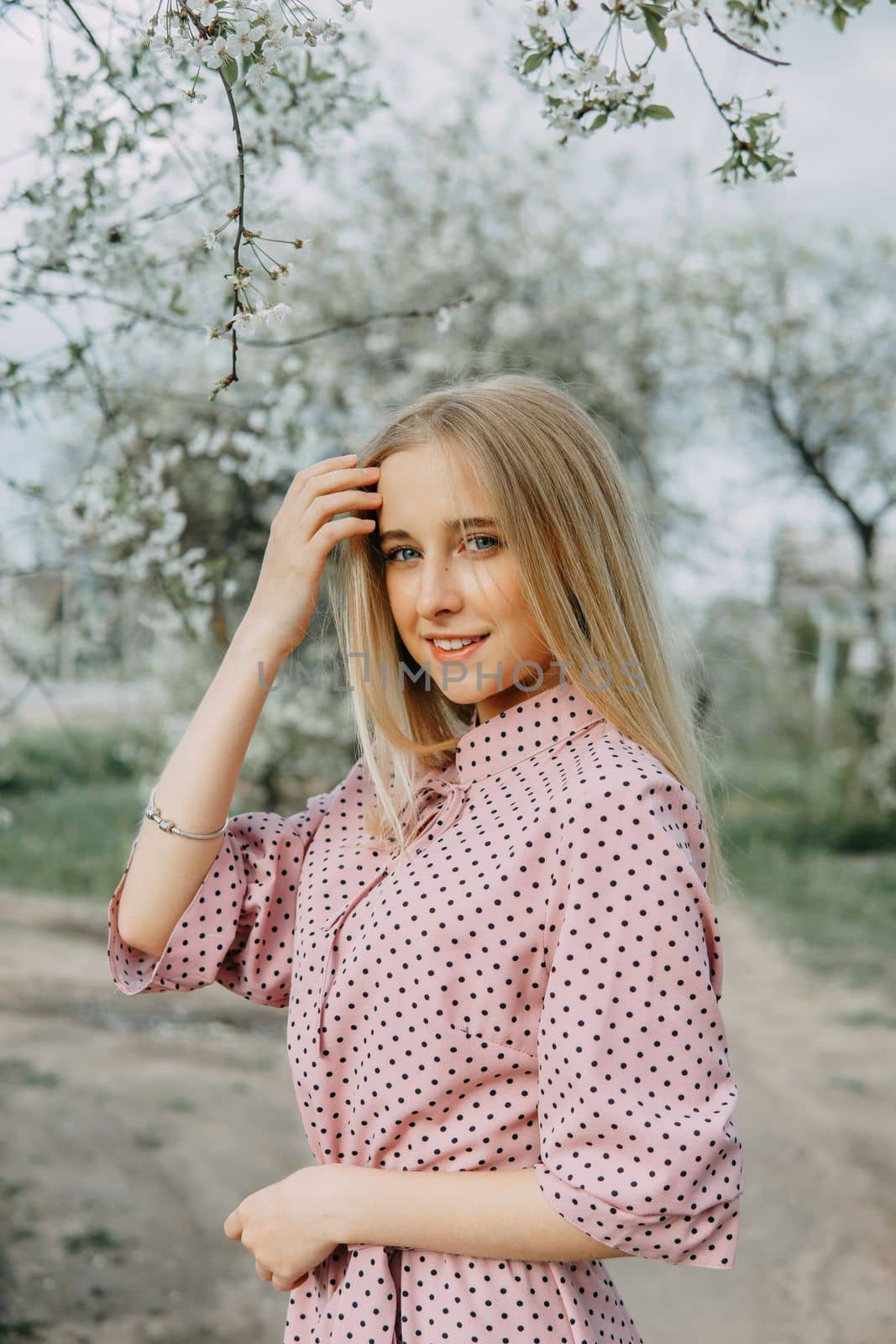 Blonde girl on a spring walk in the garden with cherry blossoms. Female portrait, close-up. A girl in a pink polka dot dress