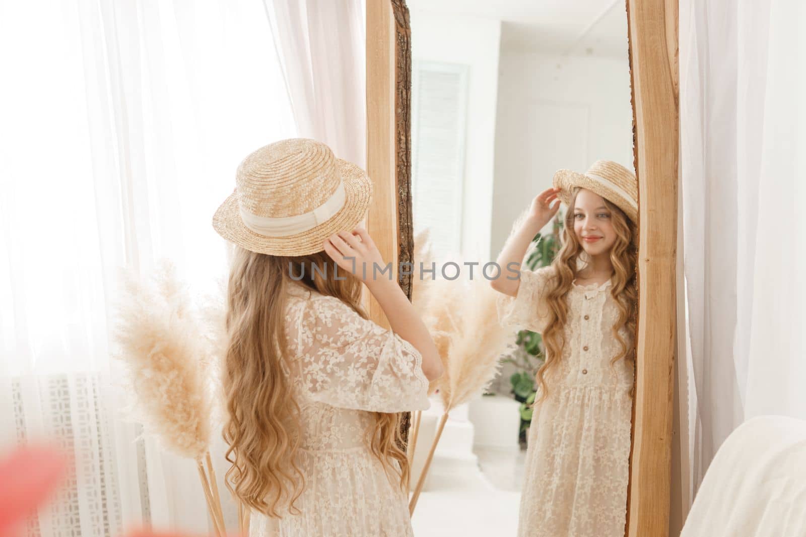 A beautiful teenage girl with long hair measures a straw hat in front of a mirror. Self-admiration of a blonde. selective focus